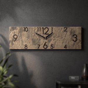 Puede incluir: Un reloj de pared rectangular de madera con acabado marrón claro. El reloj presenta números y manecillas negros. El reloj está montado en una pared gris oscuro. El reloj mide unos 61 cm de largo.