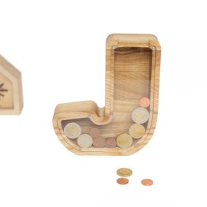 Pu&ograve; includere: Salvadanaio di legno a forma della lettera J. Il salvadanaio ha un fronte in plastica trasparente per vedere le monete all'interno. Il salvadanaio &egrave; pieno di monete.
