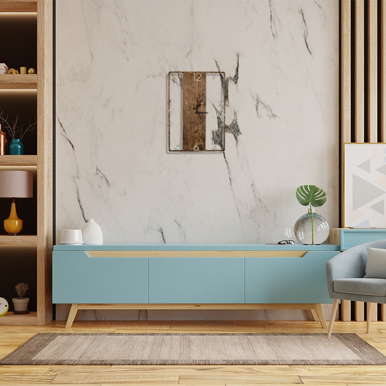 May include: A light blue media console with a wooden top and three drawers. A square wooden clock with black hands and Roman numerals is hanging on a white marble wall behind the console.