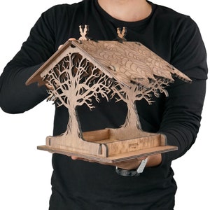Può includere: Un mangiatoia per uccelli in legno a forma di due alberi con un tetto. La mangiatoia è di colore marrone e presenta un motivo a taglio di rami. La mangiatoia è etichettata "promtl design".
