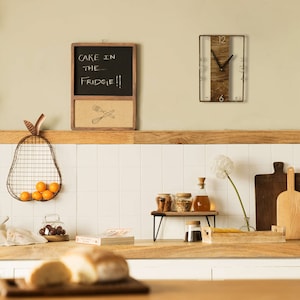 May include: A kitchen with a wooden countertop, a white tiled backsplash, and a wooden clock with a white face. The clock is hanging on the wall above a wooden shelf with a glass jar, a bottle, and a bowl of spices. There is a chalkboard with the text "Cake in the fridge!!" hanging on the wall above the shelf. There is a wire pear-shaped fruit basket with oranges on the counter.