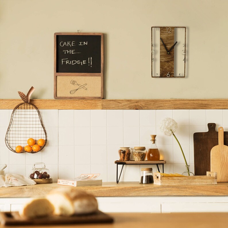 May include: A kitchen with a wooden countertop, a white tiled backsplash, and a wooden clock with a white face. The clock is hanging on the wall above a wooden shelf with a glass jar, a bottle, and a bowl of spices. There is a chalkboard with the text "Cake in the fridge!!" hanging on the wall above the shelf. There is a wire pear-shaped fruit basket with oranges on the counter.