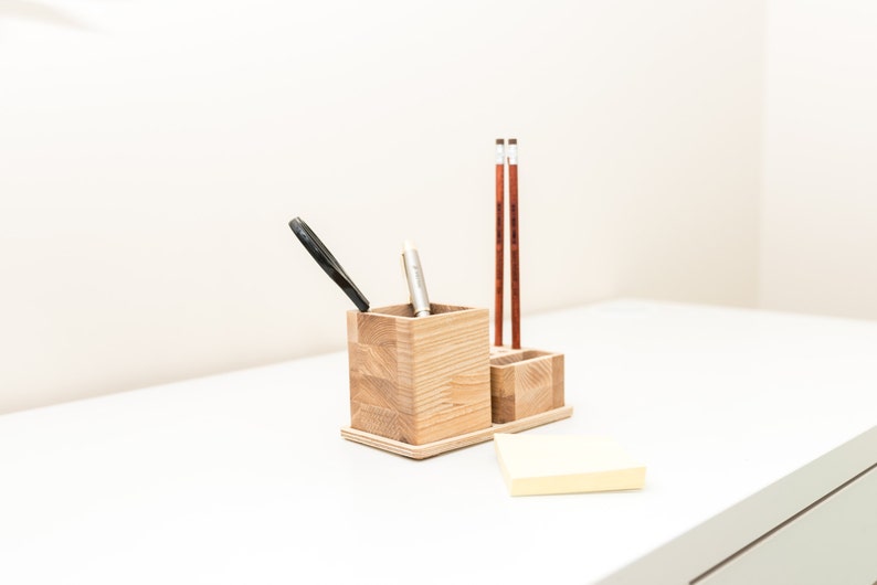 May include: A wooden desk organizer with two compartments and a small tray. The organizer holds a black pen, a silver pen, and two pencils. A yellow sticky note pad rests on the white surface.