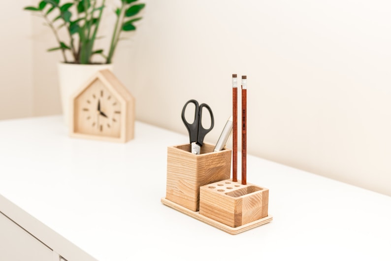 May include: A wooden desk organizer with a rectangular base, holding a square container with scissors and a pen, and a smaller section with pencil holes. Two pencils stand upright. A wooden house-shaped clock is in the background.