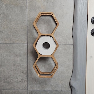 Puede incluir: Un portarrollos de papel higiénico de madera con forma de panal, montado en una pared de azulejos grises. El soporte es de madera clara y contiene un rollo de papel higiénico blanco. El diseño es moderno y minimalista, adecuado para un baño.