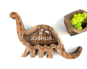 May include: Wooden dinosaur coin bank with a clear plastic window and the name "JOSHUA" engraved on the side. The bank is made of dark wood with a natural grain pattern.