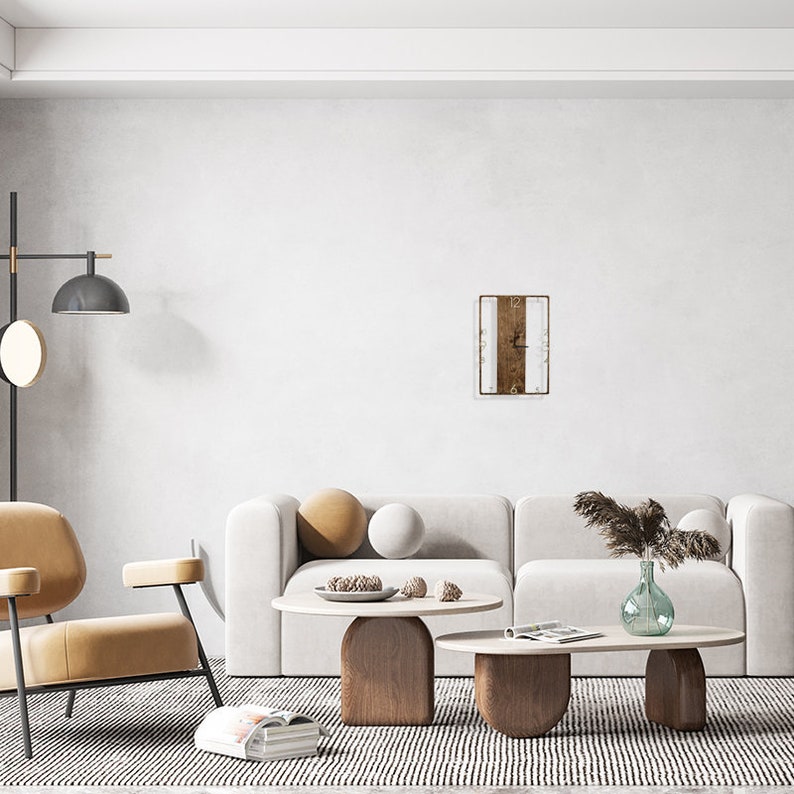 May include: A white and brown rectangular clock with Roman numerals hangs on a white wall above a white couch with two beige pillows. The couch is in a living room with a beige armchair, two wooden coffee tables, and a vase with dried grass.