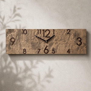 Könnte beinhalten: Eine rechteckige Wanduhr aus Holz mit natürlicher Holzmaserung. Die Uhr hat dunkelbraune Ziffern und Zeiger. Die Uhr ist an einer hellen Wand befestigt.