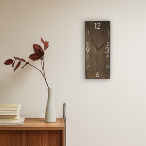 May include: A brown wooden clock with white numbers and hands. The clock is hanging on a white wall.