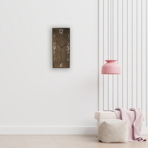 May include: A brown wooden clock with white numbers on a white wall. The clock is hanging on the wall and has a black hour hand and a black minute hand. The clock is in a room with a white lamp and a white couch.