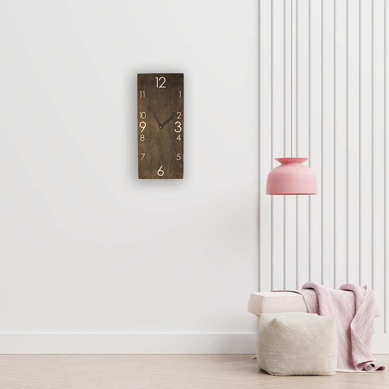 May include: A brown wooden clock with white numbers on a white wall. The clock is hanging on the wall and has a black hour hand and a black minute hand. The clock is in a room with a white lamp and a white couch.