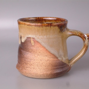 May include: A handmade ceramic mug with a brown and tan glaze. The mug has a rounded body and a slightly flared rim. The handle is attached to the side of the mug.