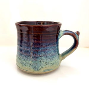 Può includere: Una tazza in ceramica fatta a mano con un'esclusiva sfumatura di colore. La tazza passa da un marrone scuro sul bordo a sfumature di blu e turchese verso la base. Ha un manico curvo e una superficie strutturata.