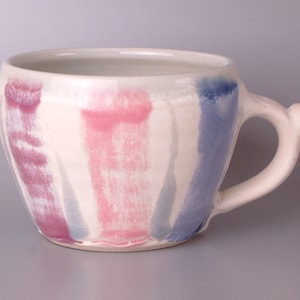 Pode incluir: Uma caneca de cerâmica branca com uma base larga e arredondada e uma alça. A caneca é decorada com listras verticais rosa, roxo e azul.