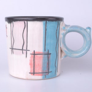 Pode incluir: Uma caneca de cerâmica com um design geométrico em tons de rosa, azul e preto. A caneca tem uma alça única.