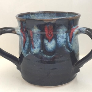 Può includere: Una tazza in ceramica fatta a mano con due manici. La tazza presenta una base blu scuro con striature rosse e azzurre. Il bordo e le maniglie sono di una tonalità di blu più scura.