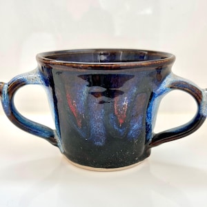 May include: A handmade ceramic mug with two handles. The mug features a dark blue glaze with streaks of lighter blue and red. The rim and handles are a darker brown color. The mug is suitable for hot or cold beverages.