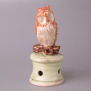 May include: A ceramic owl figurine with brown and white feathers perched on a green base with holes. The owl has closed eyes and a detailed face.