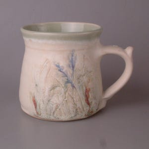 Könnte beinhalten: Eine Keramiktasse mit hellbeigem Überzug und grünem Rand. Die Tasse hat ein erhabenes Design aus Blättern und Blumen in Blautönen, Grün und Rot.