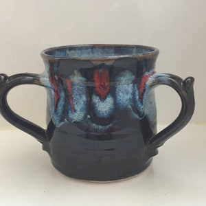 Pode incluir: Uma caneca de cerâmica artesanal com duas alças. A caneca apresenta um esmalte azul escuro com detalhes em vermelho e azul claro. As alças são de um tom de azul mais escuro e a borda tem um tom marrom.