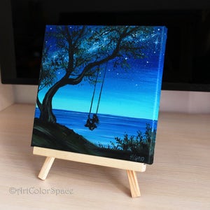 Pode incluir: Uma pintura em tela de uma silhueta de uma pessoa sentada em um balan&ccedil;o sob uma &aacute;rvore &agrave; noite. O c&eacute;u &eacute; azul escuro com estrelas brancas. O oceano est&aacute; no fundo. A pintura est&aacute; em um pequeno cavalete.