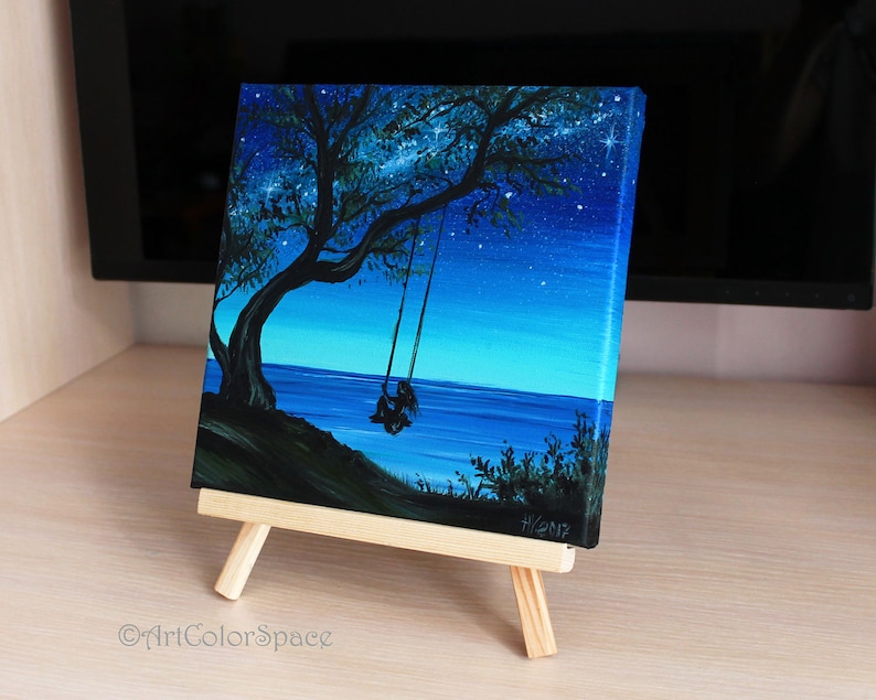 Pode incluir: Uma pintura em tela de uma silhueta de uma pessoa sentada em um balan&ccedil;o sob uma &aacute;rvore &agrave; noite. O c&eacute;u &eacute; azul escuro com estrelas brancas. O oceano est&aacute; no fundo. A pintura est&aacute; em um pequeno cavalete.