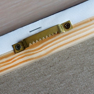 May include: A gold-colored metal hanging hook with a sawtooth edge is attached to a wooden frame with two screws. The hook is designed to hang a picture or other object on a wall.