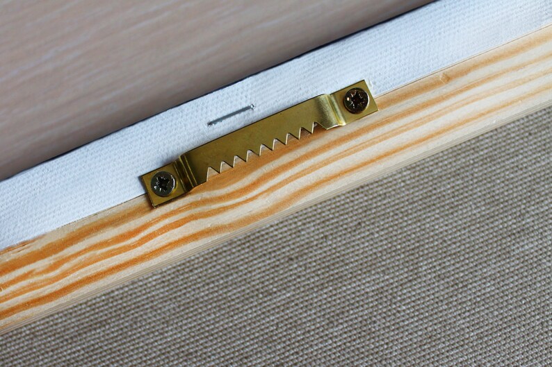 May include: A gold-colored metal hanging hook with a sawtooth edge is attached to a wooden frame with two screws. The hook is designed to hang a picture or other object on a wall.