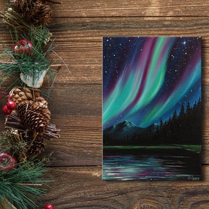 May include: A painting of the aurora borealis over a lake and forest. The sky is a deep blue with streaks of green, purple, and pink light. The lake is calm and reflective, and the forest is dark and mysterious.