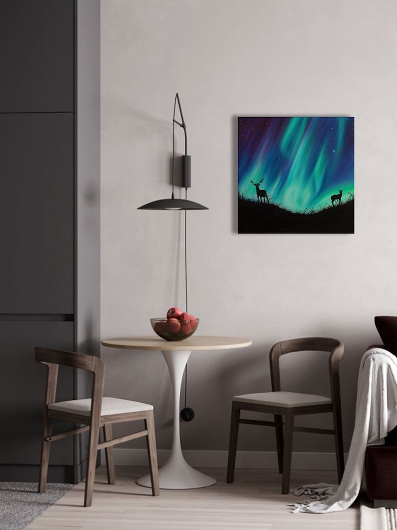 May include: A painting of a green and purple aurora borealis with two deer silhouettes in the foreground. The painting is framed and hanging on a white wall in a living room setting.