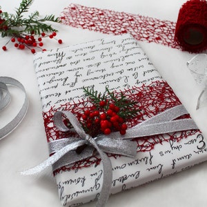 May include: A white gift box wrapped with red and silver ribbon. The box is decorated with a red mesh ribbon and a sprig of holly with red berries.