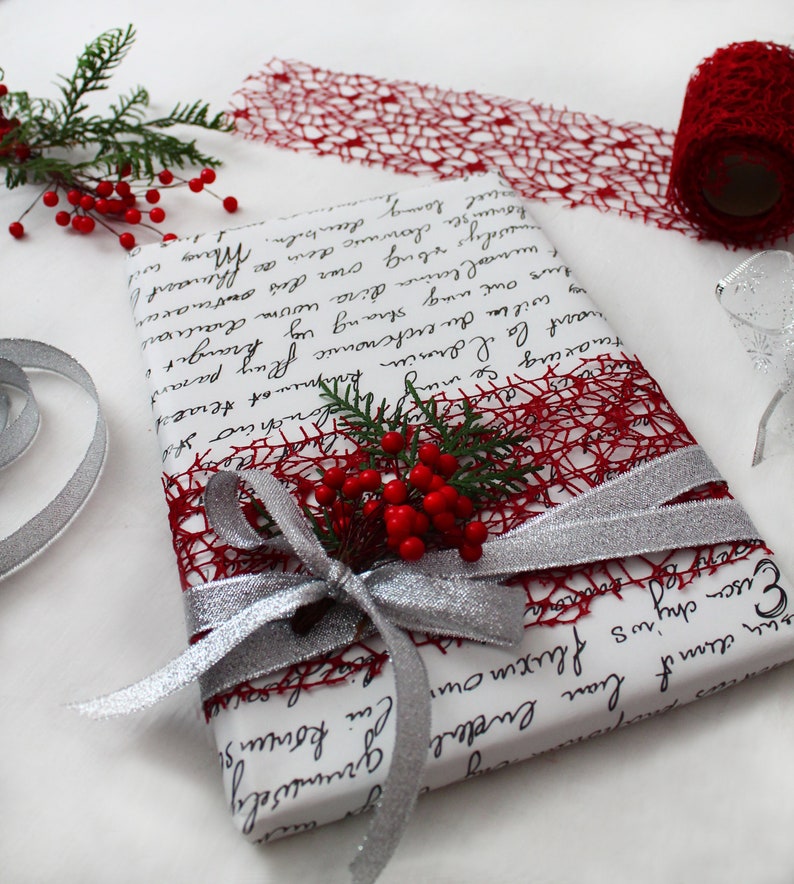 May include: A white gift box wrapped with red and silver ribbon. The box is decorated with a red mesh ribbon and a sprig of holly with red berries.