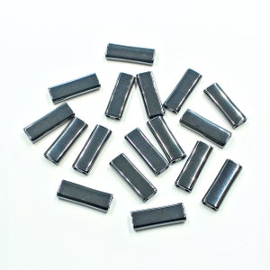 May include: A collection of rectangular, gunmetal-coloured beads with a glossy finish. Each bead has a rectangular shape with rounded edges and two small holes. The beads are scattered on a white surface.