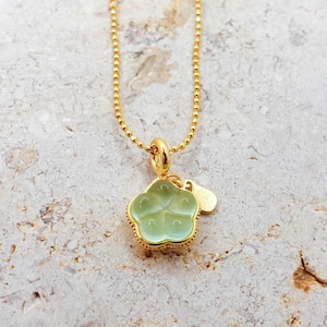 May include: A gold-colored necklace with a light green flower-shaped pendant and a small gold charm. The pendant has a detailed design and is suspended from a delicate gold chain. The necklace is displayed on a light-colored stone surface.