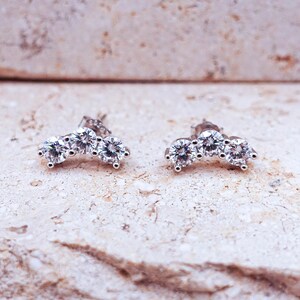 May include: A pair of silver-toned stud earrings, each featuring three clear, round-cut cubic zirconia stones arranged in an arc. The earrings are set against a light-colored, textured surface.