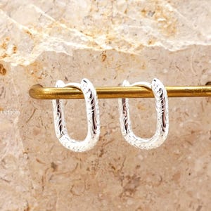 May include: A pair of silver-coloured, textured hoop earrings. The earrings are U-shaped and feature an engraved pattern. They are displayed on a gold-coloured bar against a neutral stone background. The earrings are a jewellery item.
