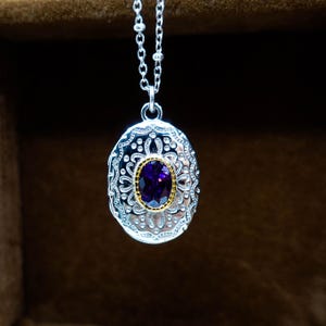 May include: Silver pendant necklace with an oval-shaped locket. The locket features an ornate design with a central purple gemstone surrounded by a gold-colored setting. The necklace chain is delicate and silver.