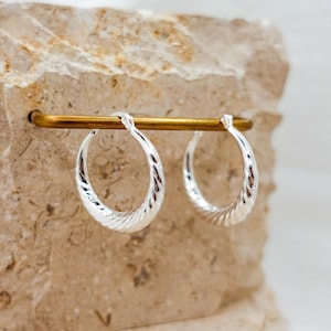 Puede incluir: Un par de pendientes de aro plateados con un diseño texturizado y acanalado. Los pendientes se exhiben en una barra dorada sobre un fondo claro y texturizado. Los aros son redondos y tienen un acabado brillante.