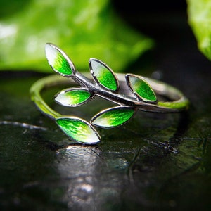 Pode incluir: Um anel de prata com um delicado design de ramo com folhas de esmalte verde. O anel est&aacute; sobre um fundo escuro.