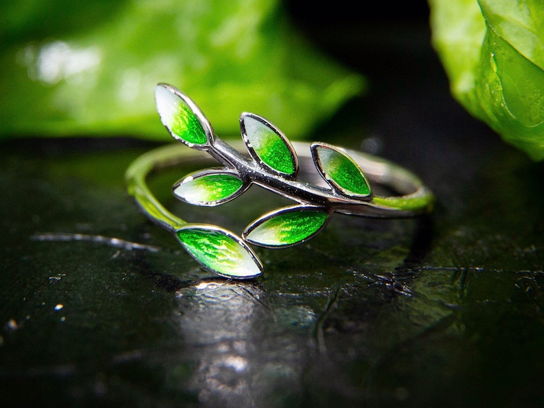 Pode incluir: Um anel de prata com um delicado design de ramo com folhas de esmalte verde. O anel est&aacute; sobre um fundo escuro.