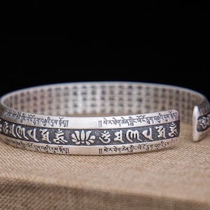 May include: Silver bracelet with a repeating pattern of Tibetan script and a lotus flower design. The bracelet is open and has a gap on one side.