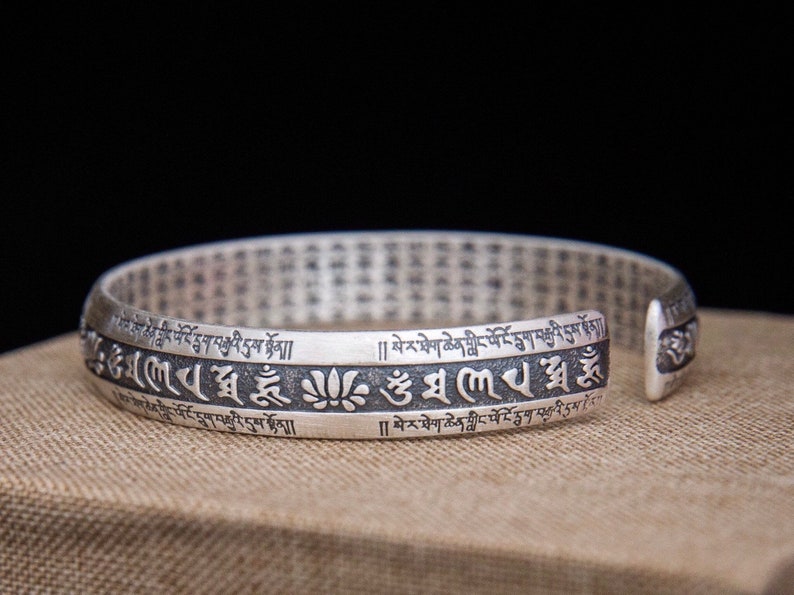 May include: Silver bracelet with a repeating pattern of Tibetan script and a lotus flower design. The bracelet is open and has a gap on one side.