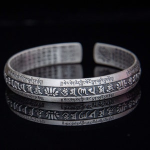 May include: A silver bracelet with a repeating pattern of Tibetan script and lotus flower designs. The bracelet is engraved with the text "][སེ་ར་ཐེག་ཆེན་གླིང་ལོ་ངོ་དྲུག་བརྒྱའི་དུས་སྟོན།།  །། སེ་ར་ཐེག་ཆེན་གླིང་པོ་ངོ་དྲུག་བརྒྱའི་དུས་མེད"