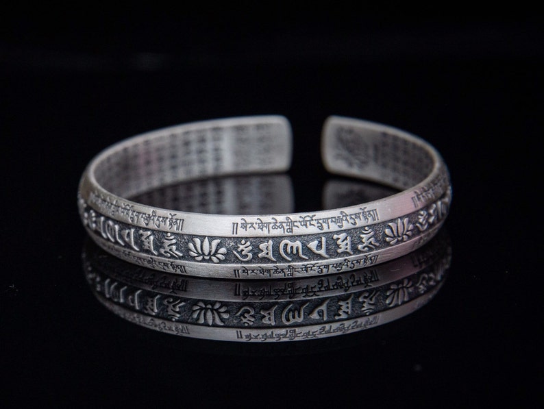 May include: A silver bracelet with a repeating pattern of Tibetan script and lotus flower designs. The bracelet is engraved with the text "][སེ་ར་ཐེག་ཆེན་གླིང་ལོ་ངོ་དྲུག་བརྒྱའི་དུས་སྟོན།།  །། སེ་ར་ཐེག་ཆེན་གླིང་པོ་ངོ་དྲུག་བརྒྱའི་དུས་མེད"
