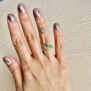 Pode incluir: Um anel de prata com um design de folha de esmalte verde. O anel &eacute; usado no dedo m&eacute;dio de uma m&atilde;o com uma tatuagem de sol.