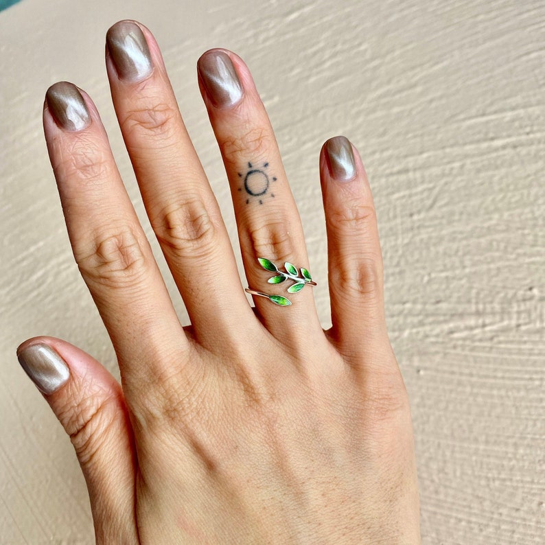 Pode incluir: Um anel de prata com um design de folha de esmalte verde. O anel &eacute; usado no dedo m&eacute;dio de uma m&atilde;o com uma tatuagem de sol.
