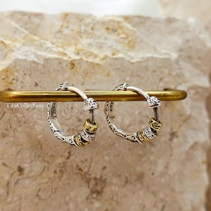 May include: A pair of silver hoop earrings with intricate etched designs and gold-colored accents. The earrings are displayed on a gold-toned bar against a neutral background. The earrings have a vintage or bohemian aesthetic.