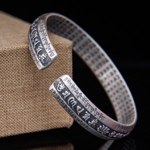 May include: A silver bracelet with Tibetan script and floral designs. The bracelet is open and adjustable.