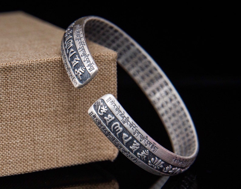May include: A silver bracelet with Tibetan script and floral designs. The bracelet is open and adjustable.