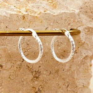 May include: Silver hoop earrings with a textured, twisted pattern. The earrings are suspended from a gold-coloured bar, set against a light-coloured stone backdrop. The brand name "TibiCollection" is visible.